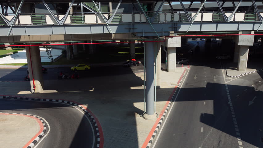 Aerial view of bustling city traffic beneath an elevated structure. Cars and motorcycles navigate curving roads under bright sunlight, showcasing urban mobility and infrastructure