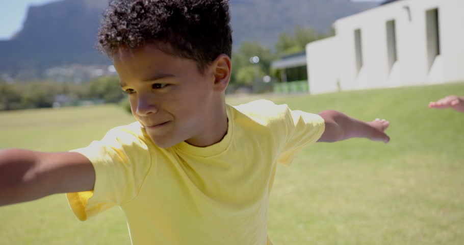 Biracial boy with curly hair is smiling outdoors in school, wearing a yellow shirt. Arms outstretched, he seems to be playing or pretending to fly in a sunny, grassy area, slow motion.