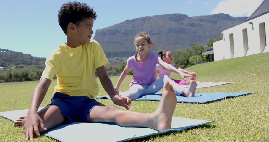 Biracial boy and girls practice yoga outdoors, with mountains in the background, in school. The boy in a yellow shirt smiles at the camera, while the girls focus on their poses, slow motion.