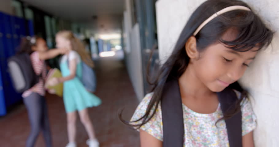 In school, girl with backpack feeling sad while other girls whispering nearby. Bullying, sadness, loneliness, childhood, emotions, unaltered, slow motion