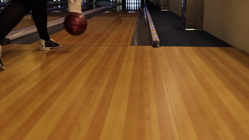 Woman plays bowling on a shiny wooden lane. The female player skillfully rolls the ball toward the pins. Team-based offline activity indoor bowling alley with bright, illuminated lanes