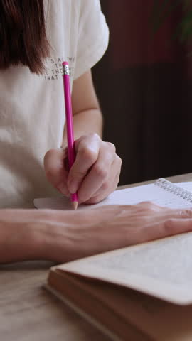 Close-up image of a brunette student preparing her lessons at an office table. The girl reads and studies from an old book and takes notes with a pencil in a notebook. The young woman studies at home