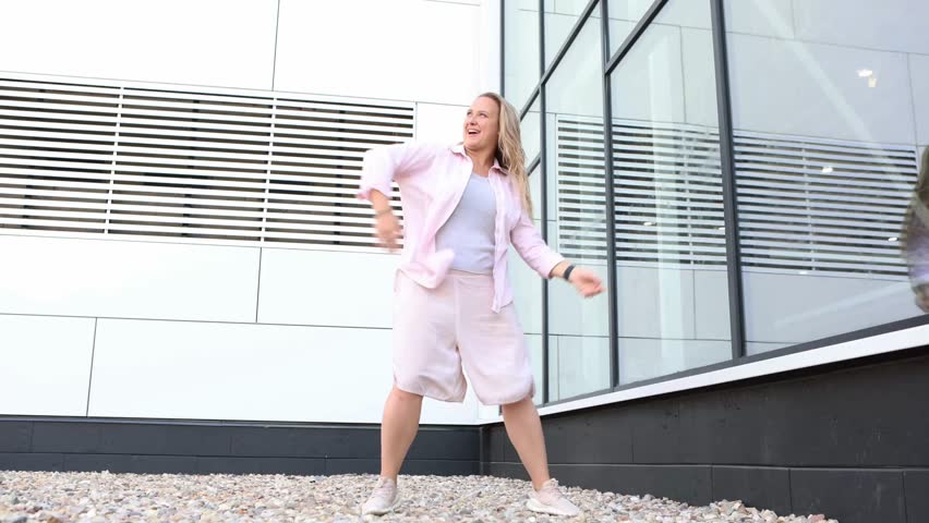 Happy Young Plus Size Woman Dancing outdoors on City Street at summer day Against Modern White Building with Glass Windows, feeling carefree and laughing with joy. Modern urban lifestyle