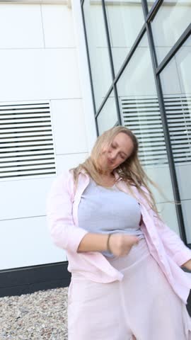 Happy Young Plus Size Woman Dancing outdoors on City Street at summer day Against Modern White Building with Glass Windows, feeling carefree and laughing with joy. Modern urban lifestyle