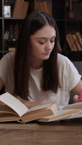 A cheerful young female student is browsing through an old book in the library and taking notes for school. A classic library and a girl enthusiastically reading specialized books for studies