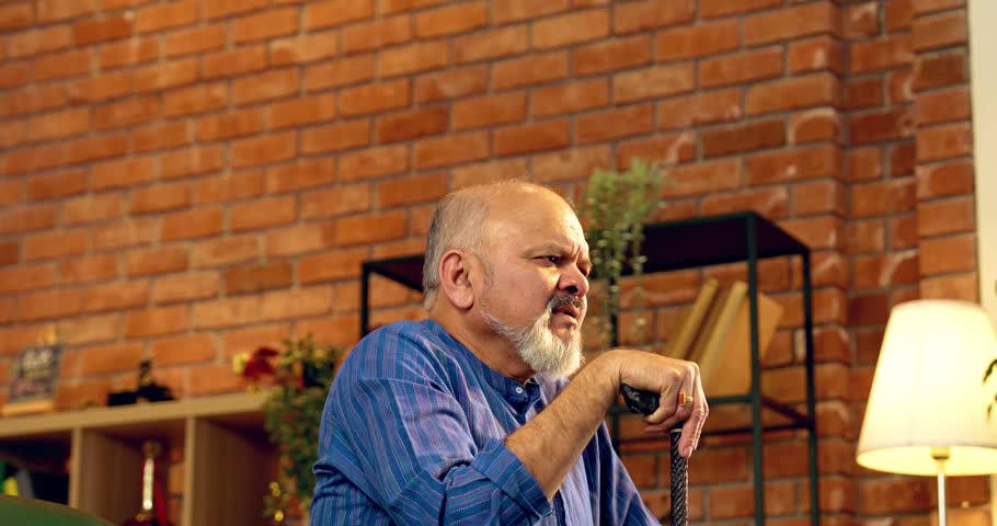 Indian elderly retired man with long beard wearing kurta, struggling to stand up with a walking stick, sitting on a sofa in a modern living room with helpless expressions and visible effort