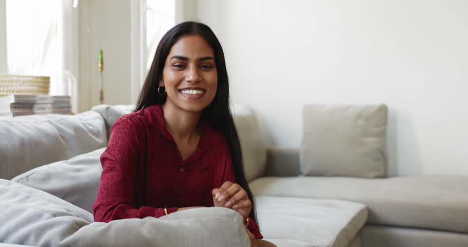 Portrait of young beautiful Indian woman looking at camera seated on sofa at home, having attractive appearance, optimistic expression, posing alone in modern living room at own or rented apartment