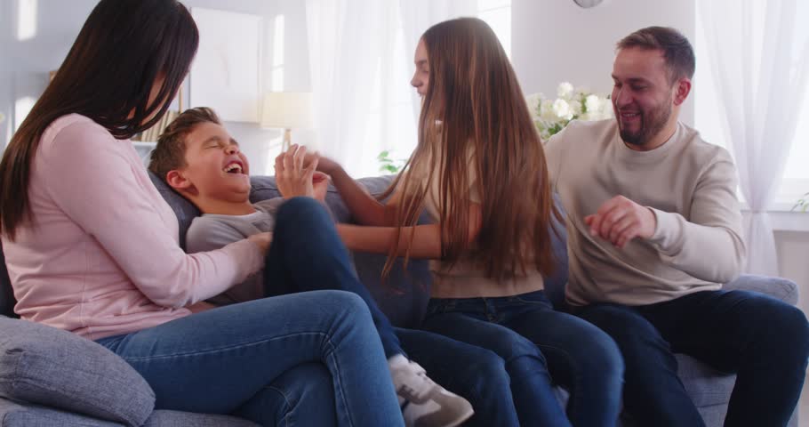 Overjoyed family have fun together tickle small boy on sofa laugh happily express positive emotions have good relationships enjoy spare time being in good mood. Family time concept