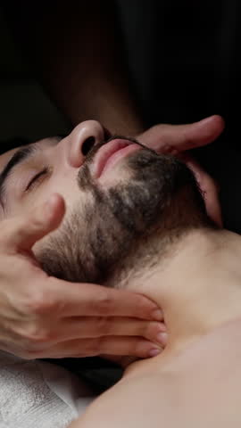Professional masseur performing slow neck massage, relieving therapeutic tension for relaxed male client lying on massage table against dark background