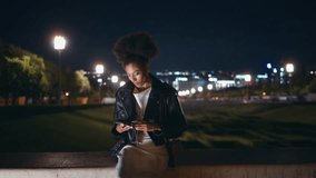 Relaxed lady messaging mobile phone sitting night street with champagne glass. Smiling african american woman reading sms on cellphone at outdoors party. Curly-haired girl relaxing at evening event. - Powered by Shutterstock - Get 15% off with code: PIKWIZARD15