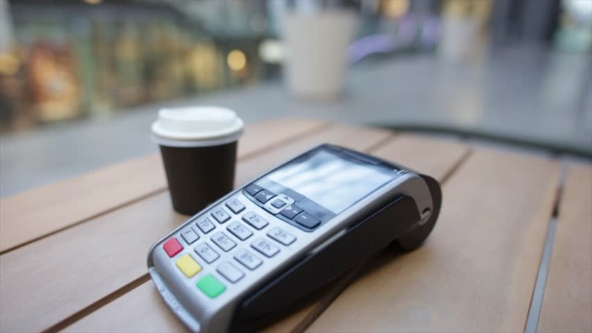 Contactless digital payment process involving customer using smartphone near point of sale terminal, completing wireless transaction beside takeaway coffee cup in contemporary coffee shop setting