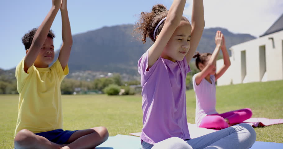 Biracial children practice yoga outdoors, with mountains in the backdrop, in school. The girl in purple leads the pose, flanked by a boy in yellow and another girl, slow motion.