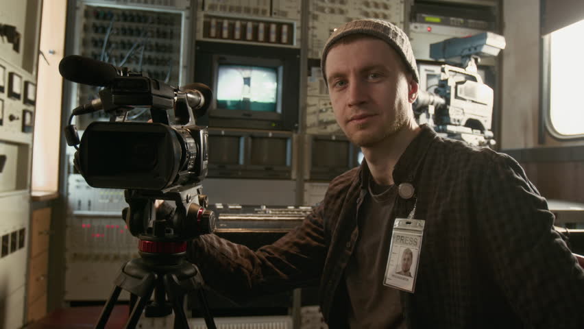 Medium closeup portrait of handsome young Caucasian male operator in beanie sitting in TV production trailer, among broadcasting equipment, with professional camera, smiling confidently