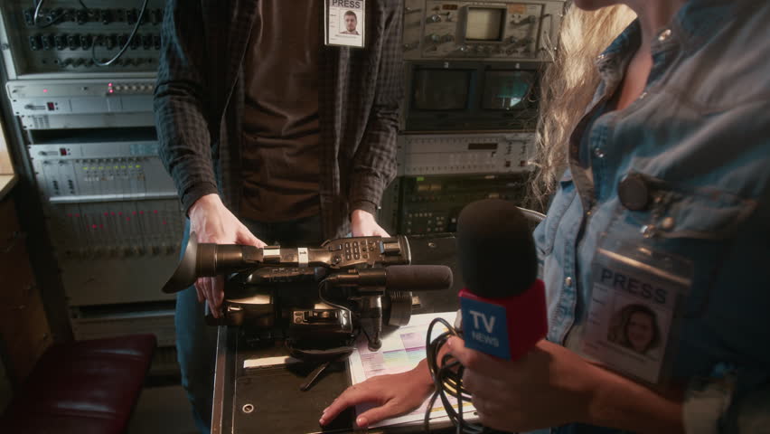 Medium full zoom-out shot of female journalist with press ID badge talking to male operator, discussing news episode timing, then taking camera and walking outside of production van to shoot in street