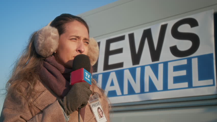 Slowmo medium closeup of face of concerned young Caucasian female journalist in earmuffs, warm coat, scarf, with press ID pass, delivering news piece on live broadcast by TV production van