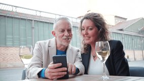 Joyful Mature Couple Sharing Special Moments While Engaging With Their Smartphone And Enjoying Delicious White Wine Together At A Cozy Table In An Outdoor Setting Filled With Warm Smiles And Laughter - Powered by Shutterstock - Get 15% off with code: PIKWIZARD15