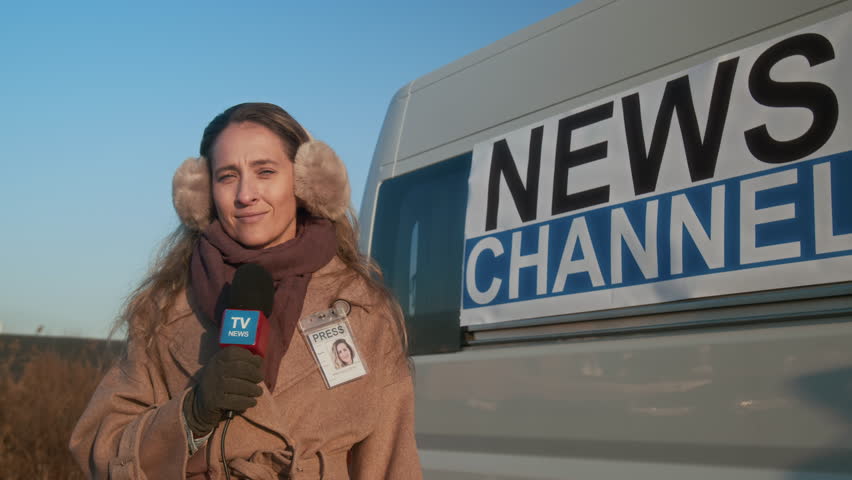 Medium portrait of cheerful young Caucasian female journalist in earmuff headphones, coat, with news channel microphone, standing outdoors on winter day, ready to broadcast news report