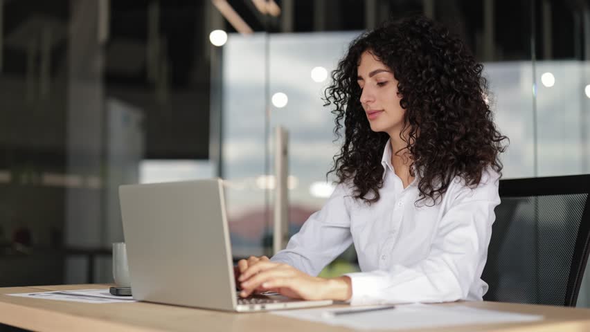 Young curly-haired businesswoman in office works on laptop. Professional woman sitting at desk with computer, successful business person using modern technology.