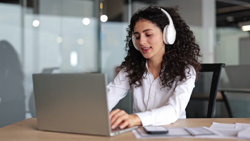 Young businesswoman with curly hair wearing headphones works on a laptop in a modern office. She types, listens, and smiles.