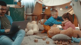 Little children play tickling each other in a make-ahead baby tent at home while their father works on the laptop, having an online business video call. - Powered by Shutterstock - Get 15% off with code: PIKWIZARD15