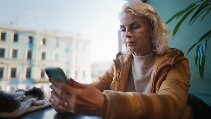 Charming 60s years-old woman resting in cafe and calling by mobile phone. Portrait of good-looking mature lady sitting alone at table with city view, aging and retirement, natural female beauty