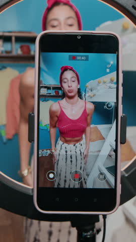 Vertical view of smartphone on tripod with ring lamp filming gen Z girl dressed in knitted top and long skirt who dancing for camera in dorm