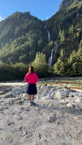 This serene video captures a young girl standing near a gently flowing river, gazing at a breathtaking waterfall in the distance. Perfect for travel documentaries, and nature-related projects.