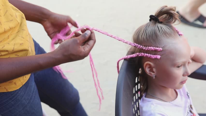 A dark-skinned woman braids a blonde girl