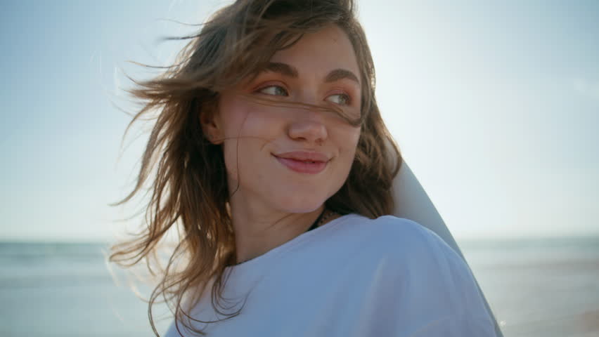 Portrait surf woman posing on ocean coast. Windy hair girl resting sea shore on sunny day. Sensual model face looking at camera laughing. Relaxed cheerful lady standing with shiny smile at sandy beach