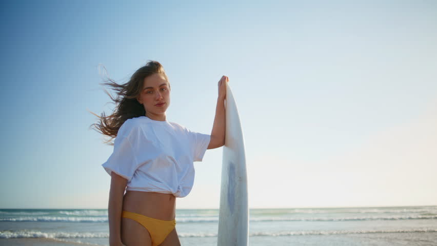 Sexy model holding surfboard at summer sandy beach closeup. Wavy haired woman surfer in bikini looking camera posing with sport equipment in marine nature. Girl enjoying light wind relaxing at ocean