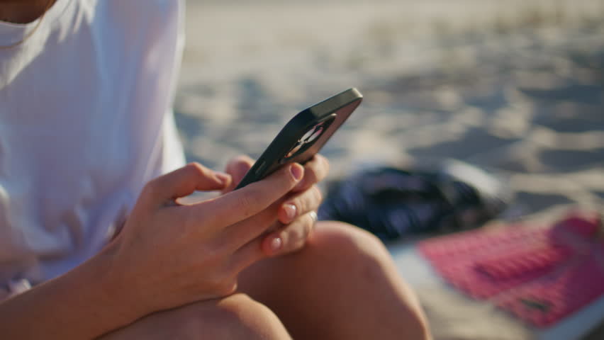 Sunny woman fingers messaging smartphone at sandy beach closeup. Smiling surprised girl texting mobile phone sms resting alone at windy ocean side. Cute emotional lady using cellphone device outdoors