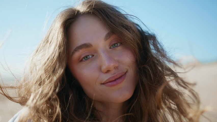 Windy hair woman face smiling on camera at blue sky horizon portrait. Young carefree girl posing on summer oceanside touching hairstyle closeup. Beautiful happy lady standing alone sunny beach nature 