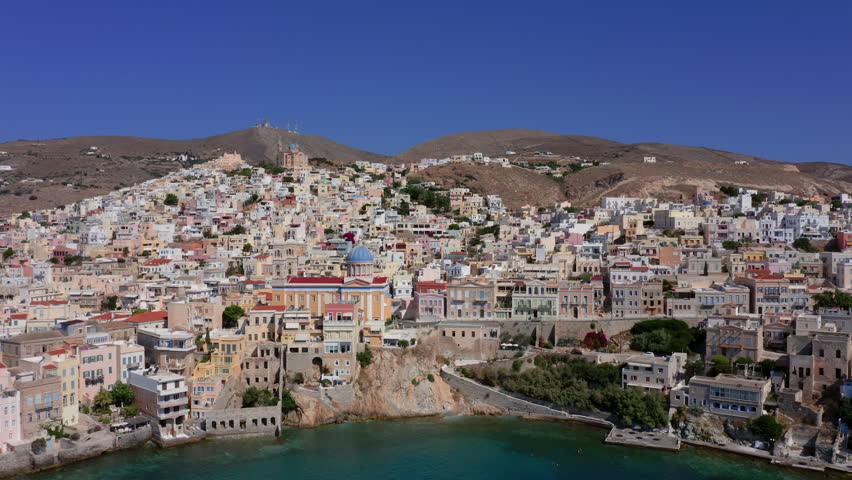 Discover an establishing panoramic aerial view of the Church of Agios Nikolaos at Asteria Beach, Syros Island, Greece. See the town sprawl across the landscape in a stunning Greek cityscape.