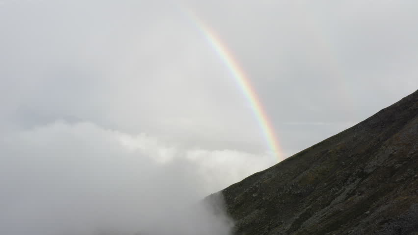 Beautiful rainbow emerging over a foggy mountain ridge, blending with the mist and rugged terrain.