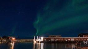 A breathtaking view of the Northern Lights (Aurora Borealis) illuminating the night sky over a Norwegian harbor. Red wooden buildings and fishing boats are brightly lit by artificial lights. - Powered by Shutterstock - Get 15% off with code: PIKWIZARD15