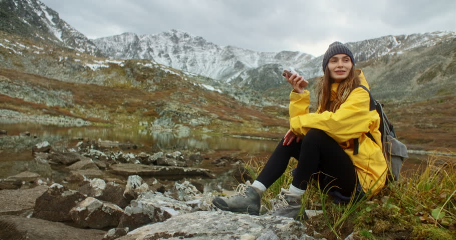 Young backpacker woman sitting by a mountain lake, using her smartphone records a voice message while soaking in the breathtaking landscape on a cloudy autumn day filled with tranquil beauty