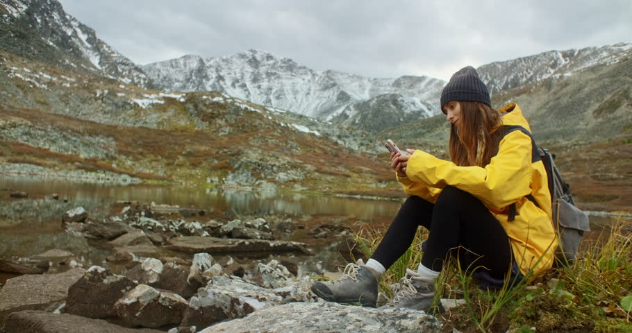 Young backpacker woman sitting by a mountain lake, using her smartphone while soaking in the breathtaking landscape on a cloudy autumn day filled with tranquil beauty