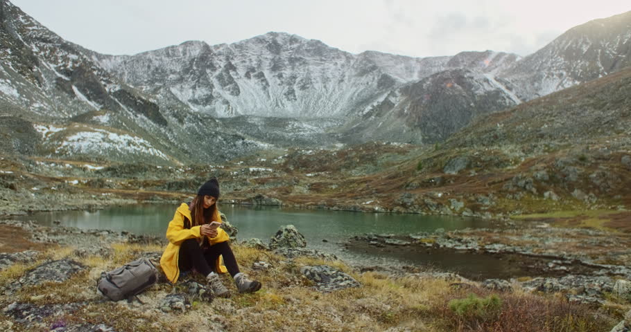 Young backpacker woman sitting by a mountain lake, using her smartphone while soaking in the breathtaking landscape on a cloudy autumn day filled with tranquil beauty