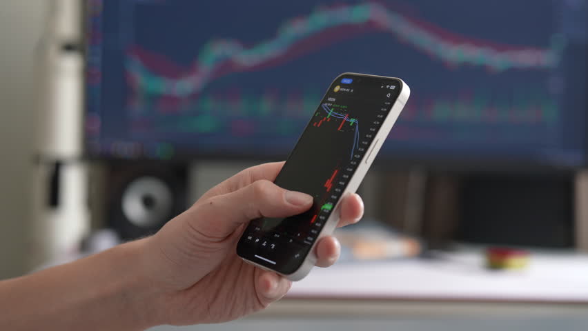 Close-up investor hand holding smartphone displaying stock market data with candlestick charts, while large monitor in background showing similar charts, suggesting active trading and market analysis.