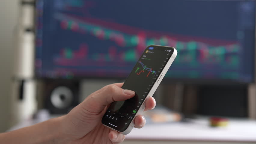 Close-up investor hand holding smartphone displaying stock market data with candlestick charts, while large monitor in background showing similar charts, suggesting active trading and market analysis. - Powered by Shutterstock - Get 15% off with code: PIKWIZARD15