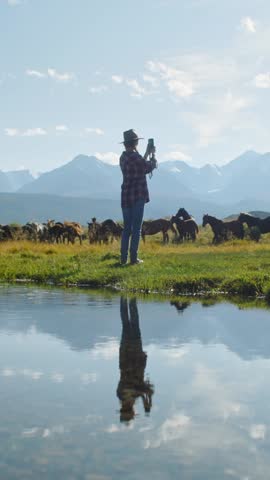 Young Woman taking pictures on phone of horses grazing in a meadow near a lake reflecting the sky and mountains