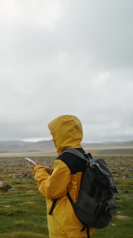 Female tourist standing hiker donned in bright yellow raincoat taking pictures on smartphone solitary tree stands tall in misty green field