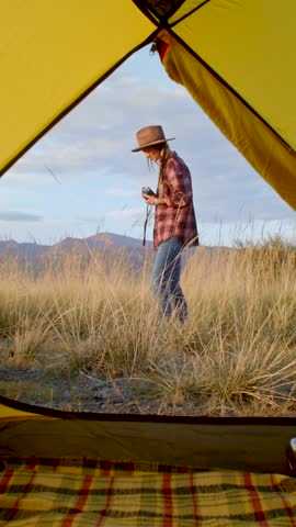 Female camper wearing hat and plaid shirt leaving yellow tent after waking up, picking up vintage film camera from blanket. Girl photographer taking pictures with vintage camera