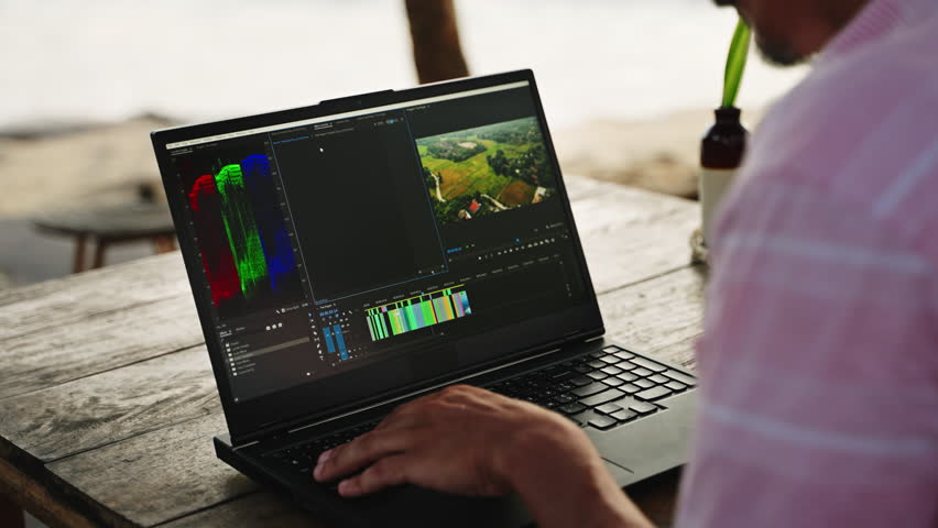 Video editor works on laptop at beachside cafe. Man uses pc for color grading clips during sunset. Freelancer edits film projects with ocean view. Creative workspace combines work, travel, relaxation.