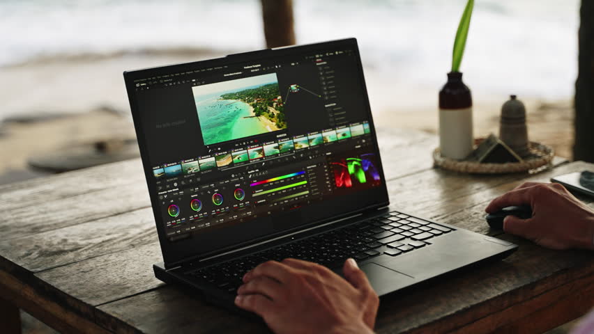 Male video editor works on laptop at beach cafe during sunset. Pro color grading footage of ocean. Freelancer edits travel film near sea on vacation. Creative workspace with view. Slow motion.