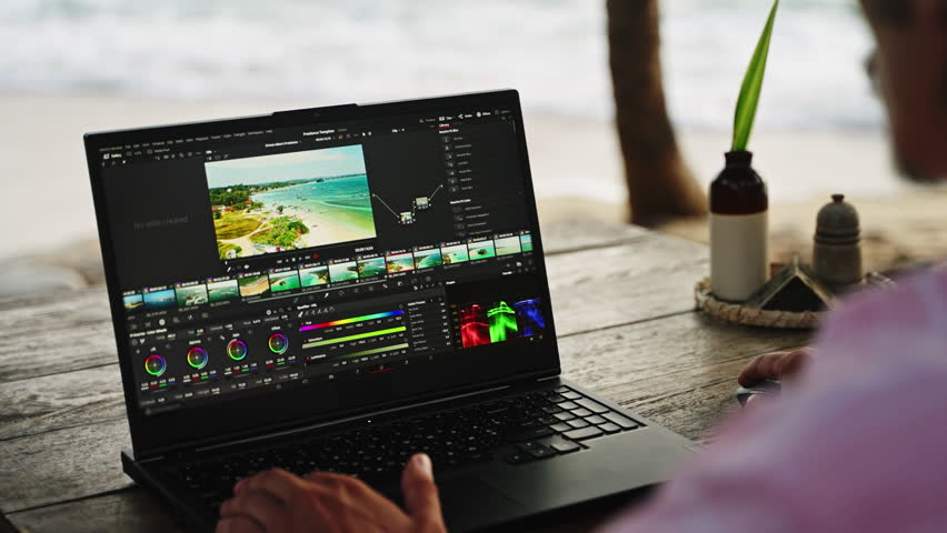 Professional video editor works on laptop with color grading tools at beach cafe, adjusting clips on timeline with ocean backdrop. Sunset lights scene as creator edits travel footage by sea. Slowmo