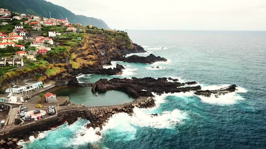Aerial view of the volcanic lava pools of Seixal, Madeira Island, Portugal, with crashing waves hitting the rocky coast and some buildings of the village on a cliff in the background