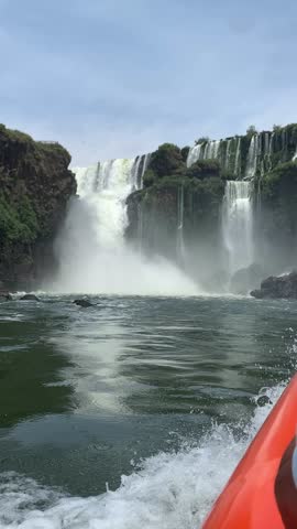 Iguazu Falls from a boat ride with cascading waterfalls, mist, rocky cliffs, and lush greenery under a bright sky. Dynamic scene showcasing the power and beauty of nature.