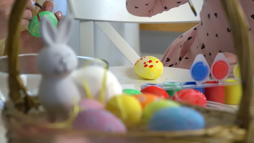 Mother and daughter painting Easter eggs together at home. Bright festive decorations, joyful atmosphere, creativity, and bonding. Colorful paints, bunny ears, and spring mood.