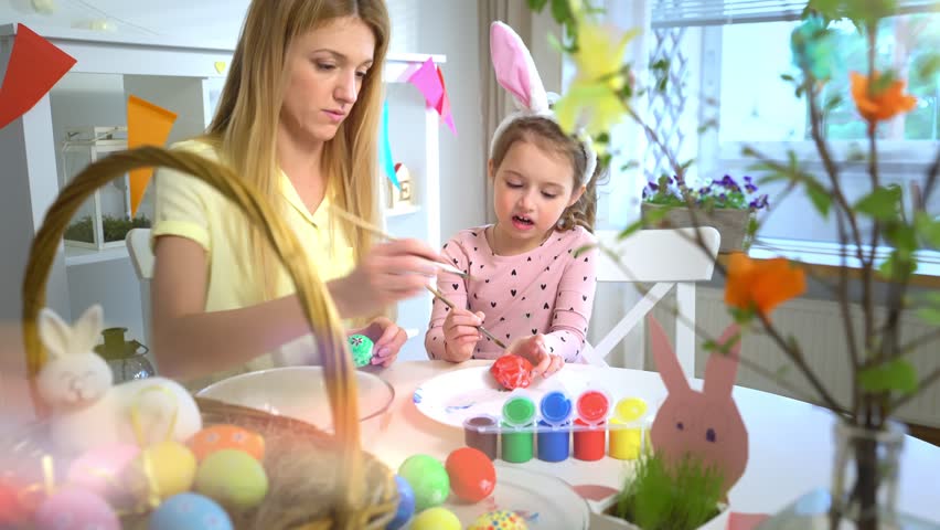 Mother and daughter painting Easter eggs together at home. Bright festive decorations, joyful atmosphere, creativity, and bonding. Colorful paints, bunny ears, and spring mood.